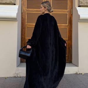 May include: A full-length black velvet coat with a flowing design. The coat is paired with a small, black handbag. The background includes a wooden door and a light-coloured wall.