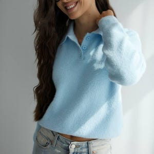 Puede incluir: Un suéter tipo polo de color azul claro con mangas largas, textura difusa, cuello y tres botones a juego en el escote, combinado con pantalones vaqueros de mezclilla clara con un botón de metal plateado y remaches cobrizos en los bolsillos.