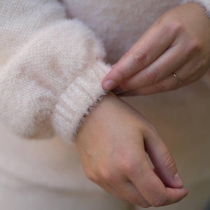 Peut inclure: Un pull à manches longues rose pâle avec une texture duveteuse et douce au toucher. Le vêtement présente un poignet en tricot côtelé au niveau de la manche, créant un effet froncé. La maille est d'une couleur blush claire uniforme.