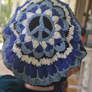 May include: A crocheted hat in shades of blue, white, and black, featuring a peace sign in the center. The hat has a round shape and a textured pattern. A small, brown tag with the text "Free From The Sky" is attached to the side.