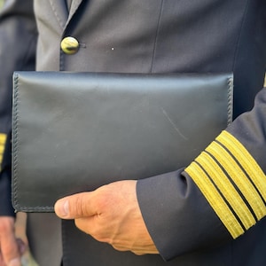 May include: A black leather wallet is held against a dark navy blue jacket. The jacket has gold stripes on the sleeve and a gold button. The wallet is rectangular and appears to be new.