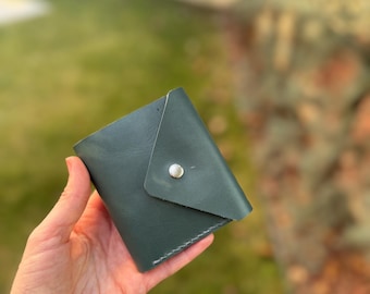 May include: A dark green leather wallet with a flap closure and a silver snap button.