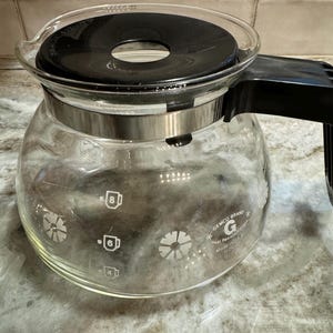 May include: A clear glass coffee carafe with a black lid and handle. The carafe has a silver band around the middle and markings for 4, 6, and 8 cups. The brand name "GEMCO BRAND" is printed on the side. Made in the U.S.A.