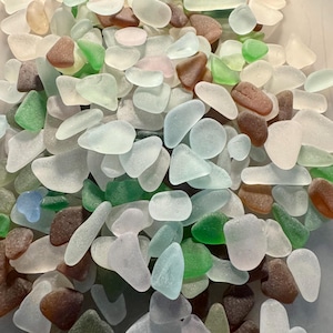 May include: A collection of sea glass in various shapes and sizes. The pieces are in shades of green, brown, white, and blue. The sea glass is smooth and frosted, with rounded edges. The pieces are scattered, creating a natural, organic look.
