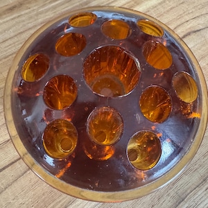 May include: A circular amber glass flower frog featuring a large central opening surrounded by twelve smaller circular holes. The translucent glass has a smooth, polished surface and a deep orange-brown hue, designed to hold flower stems upright.