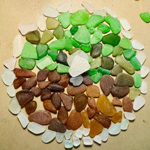 May include: A collection of sea glass pieces in various colors, including green, brown, white, and amber. The pieces are arranged in a circular pattern, showcasing their smooth, rounded shapes. The sea glass appears to be naturally tumbled and worn.