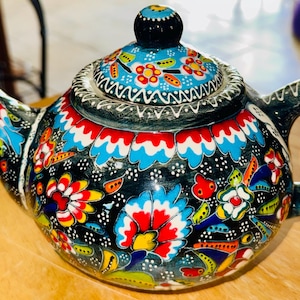 May include: A hand-painted ceramic teapot with a black base, adorned with vibrant floral patterns in red, blue, yellow, and white. The lid and handle are also decorated, creating a colorful and decorative kitchenware item.