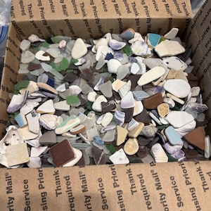 May include: A cardboard box filled with a variety of sea glass and shells. The sea glass pieces are in shades of green, blue, white, and brown. The shells are white and tan. The box has text that reads "Priority Mail Service".