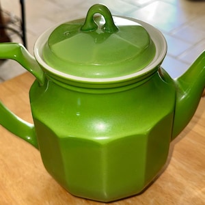 May include: A green ceramic teapot with a matching lid. The teapot has a geometric design and a curved handle. The lid has a small loop handle. The teapot is sitting on a wooden surface.