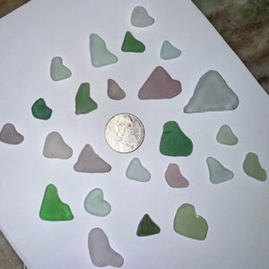 May include: A collection of twenty-four smooth, frosted sea glass pieces in various shades of green, lavender, and white, arranged around a silver-colored United States quarter coin for scale, showcasing diverse irregular shapes and rounded edges.