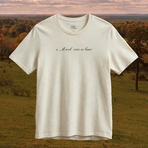 Op de afbeelding: Een off-white t-shirt met korte mouwen en de tekst "a March sister at heart" in een donker, cursief lettertype op de borst. Het shirt heeft een ronde hals, een standaard pasvorm en een klein rond logo dat aan de binnenkant van de kraag is gedrukt.