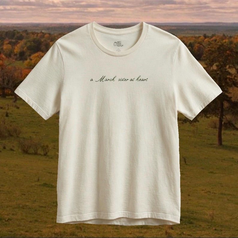 Op de afbeelding: Een off-white t-shirt met korte mouwen en de tekst "a March sister at heart" in een donker, cursief lettertype op de borst. Het shirt heeft een ronde hals, een standaard pasvorm en een klein rond logo dat aan de binnenkant van de kraag is gedrukt.