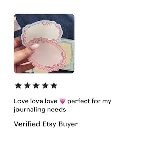 May include: A hand holding two oval-shaped, decorative paper labels with a pink and red border. The labels have a light pink center and a scalloped edge. Additional labels with blue and yellow borders are visible.