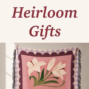 May include: A woven throw blanket featuring a pink rectangular center with white lily illustrations and the name Belle in script. The blanket has a dark scalloped border, a light purple and white gingham edge, and frayed fringe on the ends.