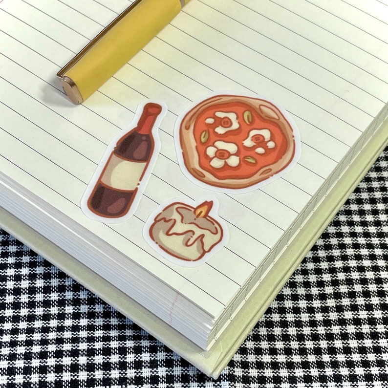 Puede incluir: Tres pegatinas en un cuaderno de l&iacute;neas: una botella de vino, una vela encendida y una pizza con flores blancas y hojas verdes.