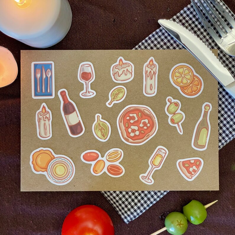Puede incluir: Una hoja de pegatinas con varios dise&ntilde;os, incluyendo copas de vino, velas, pizza, aceitunas y una botella de vino. Las pegatinas est&aacute;n sobre un fondo marr&oacute;n con un patr&oacute;n a cuadros.