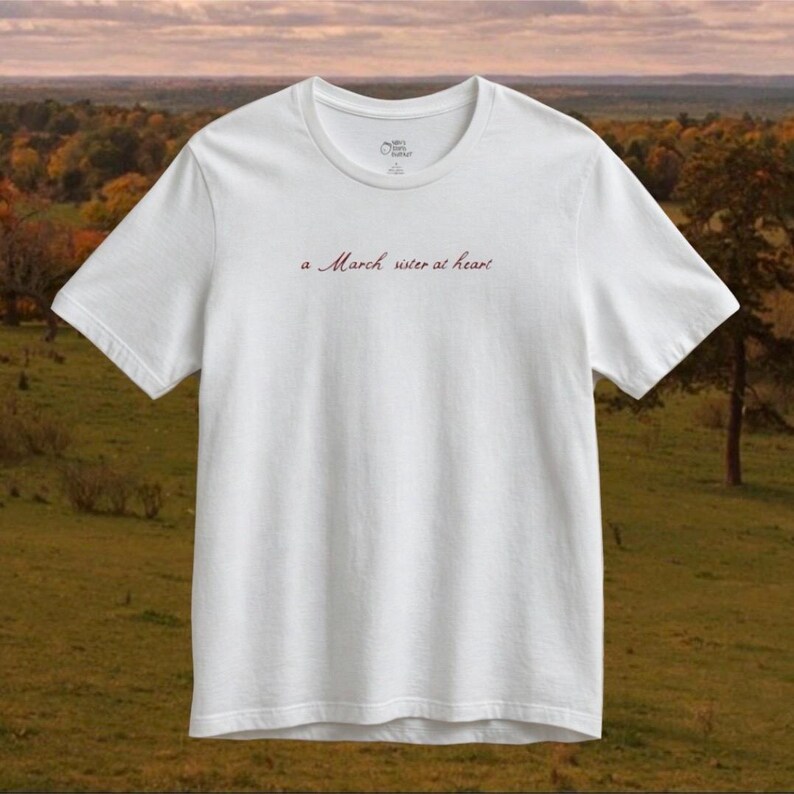Op de afbeelding: Een wit T-shirt met korte mouwen en de tekst "a March sister at heart" in een donkerrood cursief lettertype op het midden van de borst. Het shirt heeft een ronde hals en een standaard pasvorm, met een klein logo aan de binnenkant van de kraag.