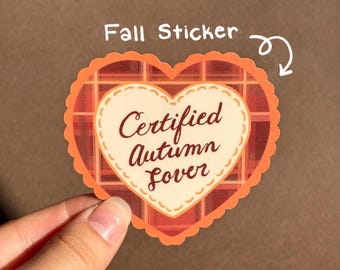 May include: A heart-shaped sticker with a scalloped orange border and a red and orange plaid pattern. The center features a cream-colored heart with a dashed border and the brown text "Certified Autumn Lover" written in a cursive script.