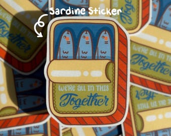Puede incluir: Una pegatina rectangular con una ilustración estilizada de una lata de sardinas abierta con tres peces azules dentro. La lata tiene un borde a rayas rojas y una etiqueta verde con el texto "WE'RE ALL IN THIS Together" en azul y blanco.