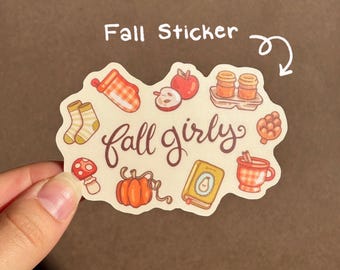May include: A die-cut sticker featuring the text fall girly in brown script surrounded by illustrations of an orange plaid oven mitt, a red apple, two coffee cups, socks, a mushroom, a pumpkin, a yellow book, and a plaid mug with cinnamon.