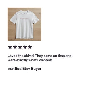 Op de afbeelding: Een wit T-shirt met korte mouwen, een ronde hals en een kleine, donkerrode cursieve tekst op de bovenkant van de borst. De tekst op het shirt luidt "A March wine at last". Het kledingstuk ligt plat op een lichte houten ondergrond.
