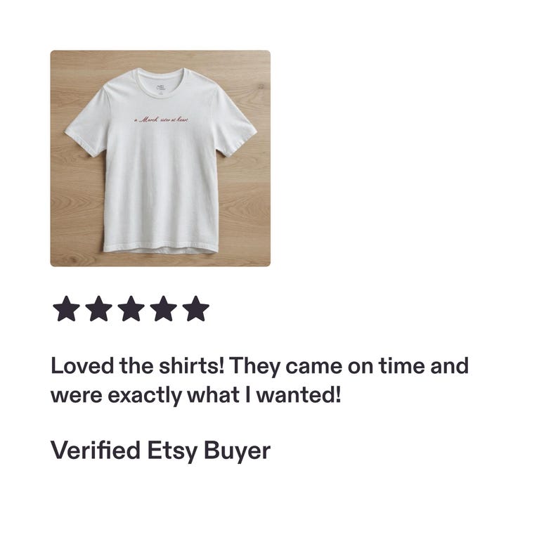 Op de afbeelding: Een wit T-shirt met korte mouwen, een ronde hals en een kleine, donkerrode cursieve tekst op de bovenkant van de borst. De tekst op het shirt luidt "A March wine at last". Het kledingstuk ligt plat op een lichte houten ondergrond.