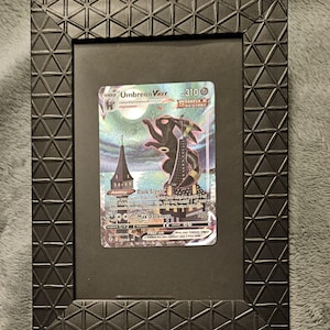 Puede incluir: Una tarjeta coleccionable enmarcada con el personaje Umbreon V. La tarjeta está colocada sobre un fondo negro dentro de un marco negro con un patrón geométrico. El arte de la tarjeta representa una criatura oscura con un telón de fondo de ciudad.