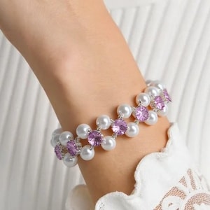 May include: A delicate bracelet featuring alternating white pearls and small, round, light purple gemstones. The bracelet is worn on a wrist, against a white ribbed long-sleeve shirt with a lace cuff.