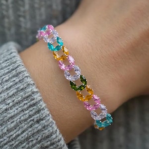 May include: A colorful bracelet with oval-shaped gemstones in shades of pink, blue, yellow, and green. The bracelet is worn on a wrist, with a gray sweater in the background. The gemstones are linked together in a chain.