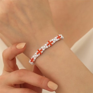 May include: A delicate bracelet featuring a floral design. The bracelet has a silver band with small white pearls and vibrant orange flower accents. The bracelet is worn on a wrist, showcasing its intricate details and elegant style.