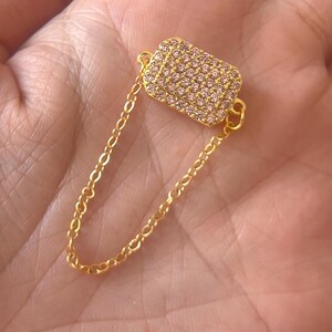 Könnte beinhalten: Ein goldfarbener Ohrschmuck mit rechteckigem Design, besetzt mit kleinen, rosa Edelsteinen. Eine zarte Goldkette verbindet das rechteckige Stück mit einer kleinen Schlaufe. Der Ohrschmuck wird vor einem hautfarbenen Hintergrund gezeigt.