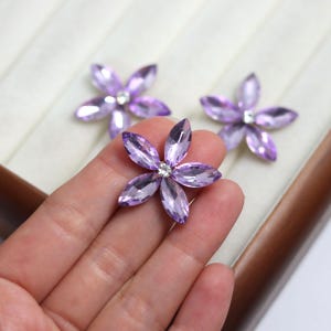 May include: Three purple flower-shaped crystal embellishments with a central clear crystal. The petals are faceted, and the crystals are set on a light-colored surface. The flowers are likely for crafts or jewelry making.