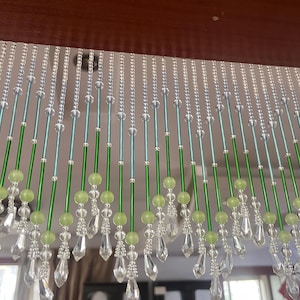 May include: A beaded curtain featuring vertical strands of clear crystal-like beads, light green spherical beads, and long green cylindrical rods. The strands terminate in clear, faceted teardrop-shaped pendants that hang at varying lengths.