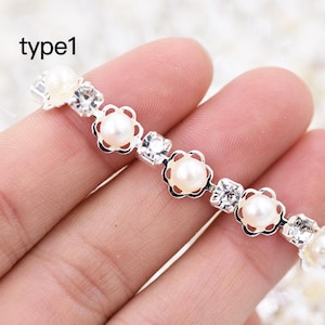 May include: A silver chain with white pearl and rhinestone accents. The chain is made of small flower-shaped links.