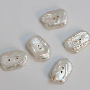 May include: Five irregular, off-white pearl buttons with two holes each. The buttons have a lustrous, iridescent sheen and a slightly textured surface. The buttons are arranged on a white background.