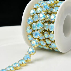 May include: A reel of heart-shaped, iridescent blue rhinestones with gold-coloured metal settings. The rhinestones are arranged in a continuous line, creating a decorative trim. The reel is white, and the background is a soft white.