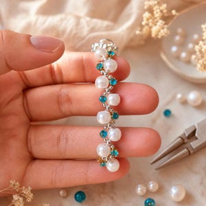 May include: A silver earring with a vertical design. The earring features alternating white pearls and teal-colored gemstones. The design is accented with small gold-colored details. The earring is held in a hand.