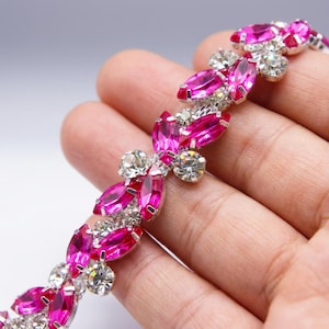 Puede incluir: Primer plano de un adorno decorativo plateado con pedrería de cristal rosa y transparente. El adorno presenta pedrería rosa en forma de marquesa y pedrería redonda transparente, dispuesta en un patrón floral. El adorno se sostiene en una mano.