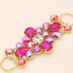 May include: A gold-toned jewelry piece featuring an arrangement of vibrant, faceted gemstones. The gemstones are primarily hot pink and iridescent, with a central clear stone. Small clear crystals and gold rings add detail.