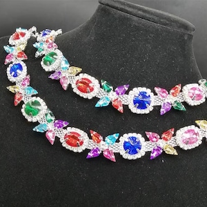 May include: Two strands of colorful rhinestone trim. The trim features a silver mesh base with a repeating pattern of oval and round rhinestones in various colors, including red, blue, green, and purple. The rhinestones are surrounded by smaller clear stones.