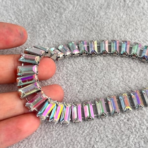 May include: A close-up of a necklace with iridescent rectangular crystals set in a silver-tone metal. The crystals reflect a rainbow of colours, including pink, yellow, and blue. The necklace is on a grey surface.