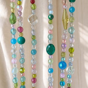 May include: Multiple vertical strands of decorative beads in various shapes, sizes, and colors including clear, light blue, lime green, pink, magenta, and teal. The beads feature faceted, smooth, and iridescent finishes, creating a colorful display.