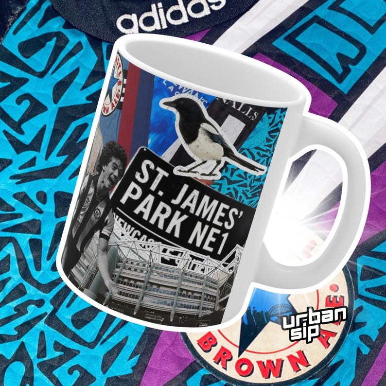 May include: White ceramic mug with a colorful collage design featuring a black and white magpie, a sign that reads "St. James' Park NE1", and a view of a stadium. The mug has a black handle.
