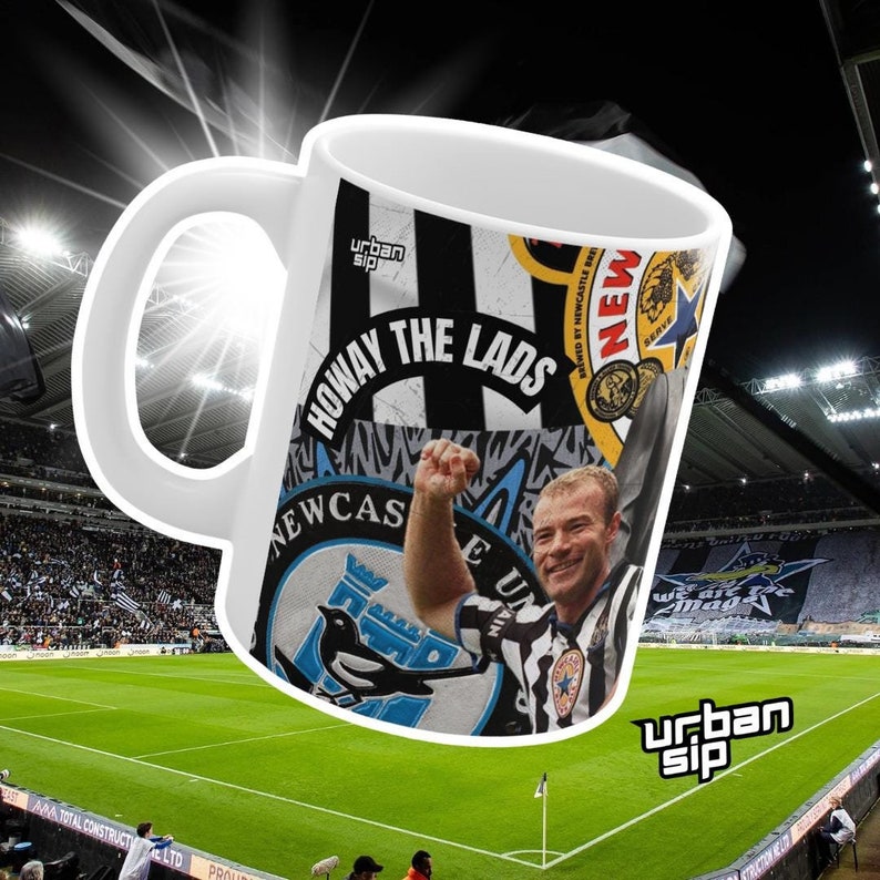 May include: White ceramic mug with a black and white design featuring a soccer player, a team logo, and the text "Howay the Lads". The mug is in front of a green soccer field with a stadium in the background.