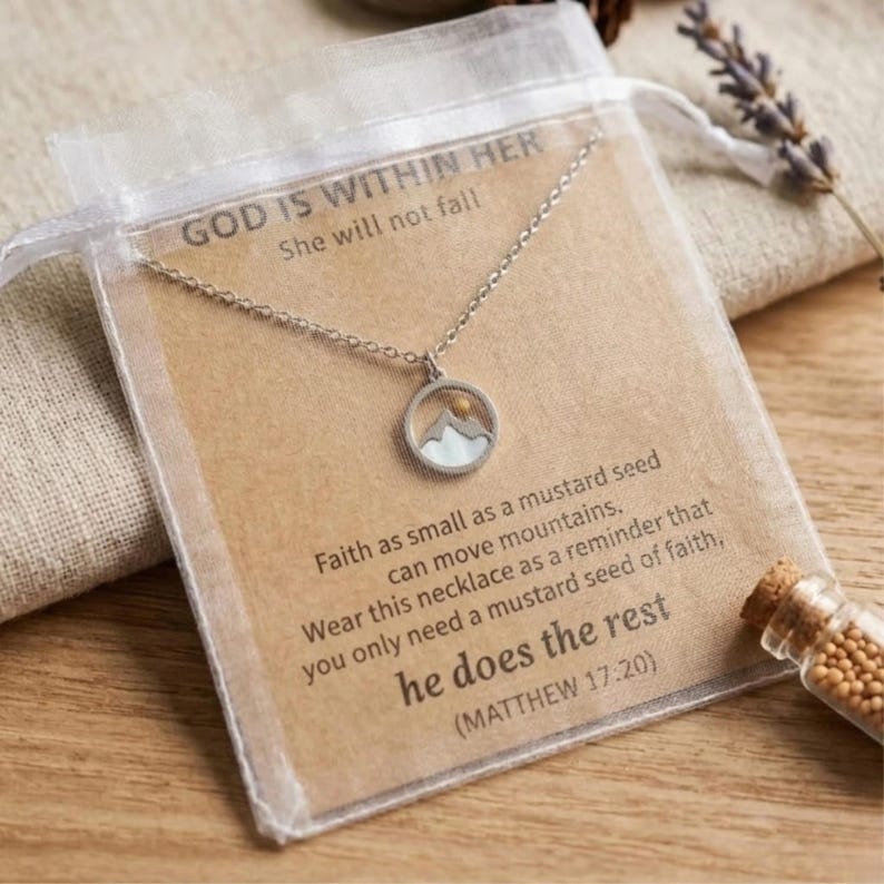 Puede incluir: Un collar plateado con un colgante de monta&ntilde;a, presentado en una tarjeta dentro de una bolsa transparente. La tarjeta tiene el texto "GOD IS WITHIN HER" y una cita de Mateo 17:20. El collar est&aacute; sobre una superficie de madera.
