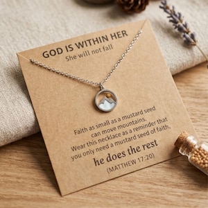 Puede incluir: Collar plateado con un colgante redondo que presenta una escena de monta&ntilde;a y un peque&ntilde;o punto dorado. El collar se muestra en una tarjeta marr&oacute;n con el texto "GOD IS WITHIN HER" y un vers&iacute;culo b&iacute;blico sobre la fe.