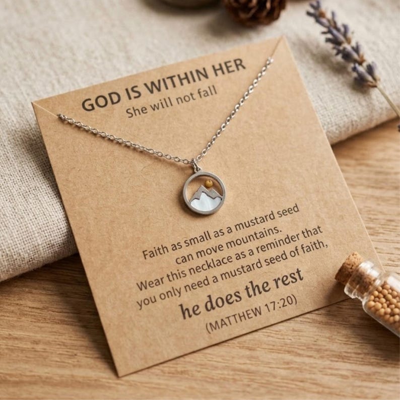 Puede incluir: Collar plateado con un colgante redondo que presenta una escena de monta&ntilde;a y un peque&ntilde;o punto dorado. El collar se muestra en una tarjeta marr&oacute;n con el texto "GOD IS WITHIN HER" y un vers&iacute;culo b&iacute;blico sobre la fe.