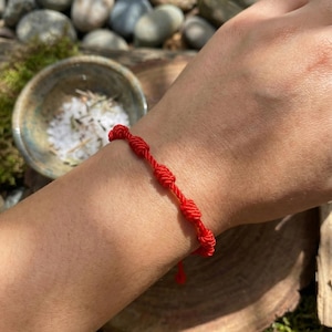 May include: A red string bracelet with multiple knots. The bracelet is worn on a person's wrist. A small ceramic bowl with white salt and herbs is in the background.