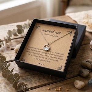 Puede incluir: Un collar plateado con un colgante de monta&ntilde;a, presentado en una caja de regalo negra. La caja contiene una tarjeta con el texto "mustard seed - the seed of faith" y texto inspirador. El collar est&aacute; sobre una superficie de madera.