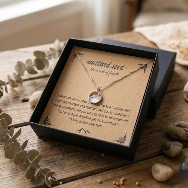 Puede incluir: Un collar plateado con un colgante de monta&ntilde;a, presentado en una caja de regalo negra. La caja contiene una tarjeta con el texto "mustard seed - the seed of faith" y texto inspirador. El collar est&aacute; sobre una superficie de madera.